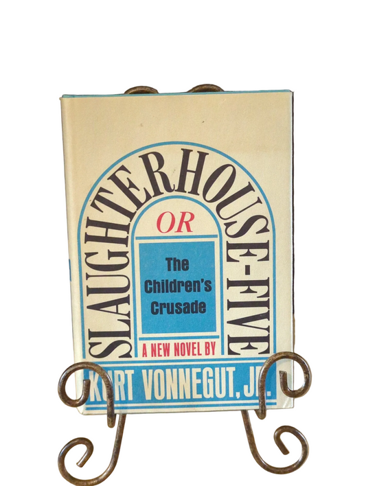 Front dust jacket of Kurt Vonnegut Jr.’s Slaughterhouse-Five on a curled metal display stand, with blue and red lettering and visible jacket toning.