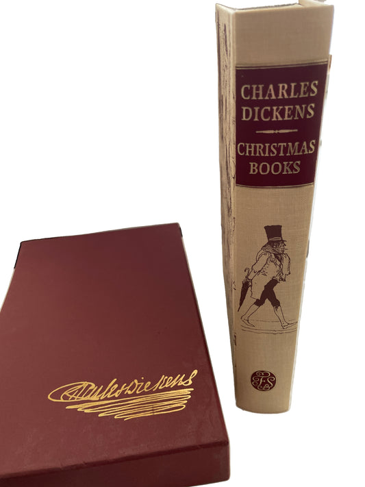 Christmas Books by Charles Dickens standing beside its burgundy slipcase, showing the cream illustrated spine and gold facsimile signature design on the case.