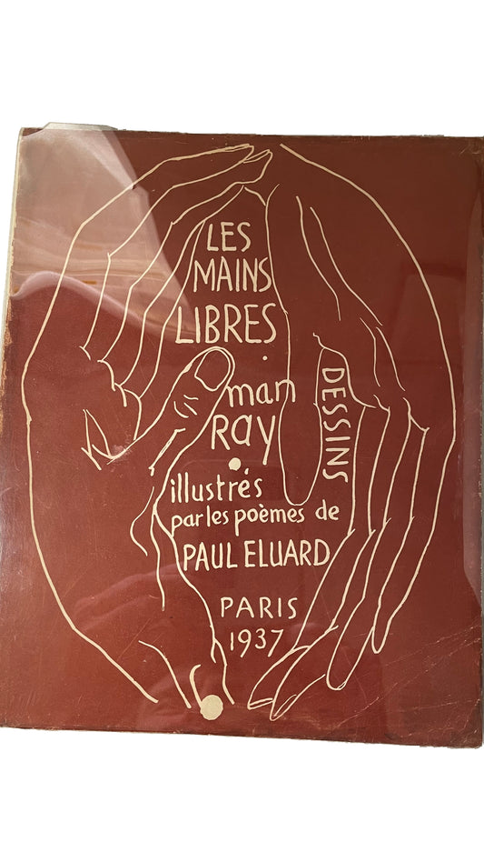 Red-brown Les Mains Libres cover by Man Ray and Paul Éluard, with cream outlined hands and lettering beneath a clear protective covering, showing reflections and wear.